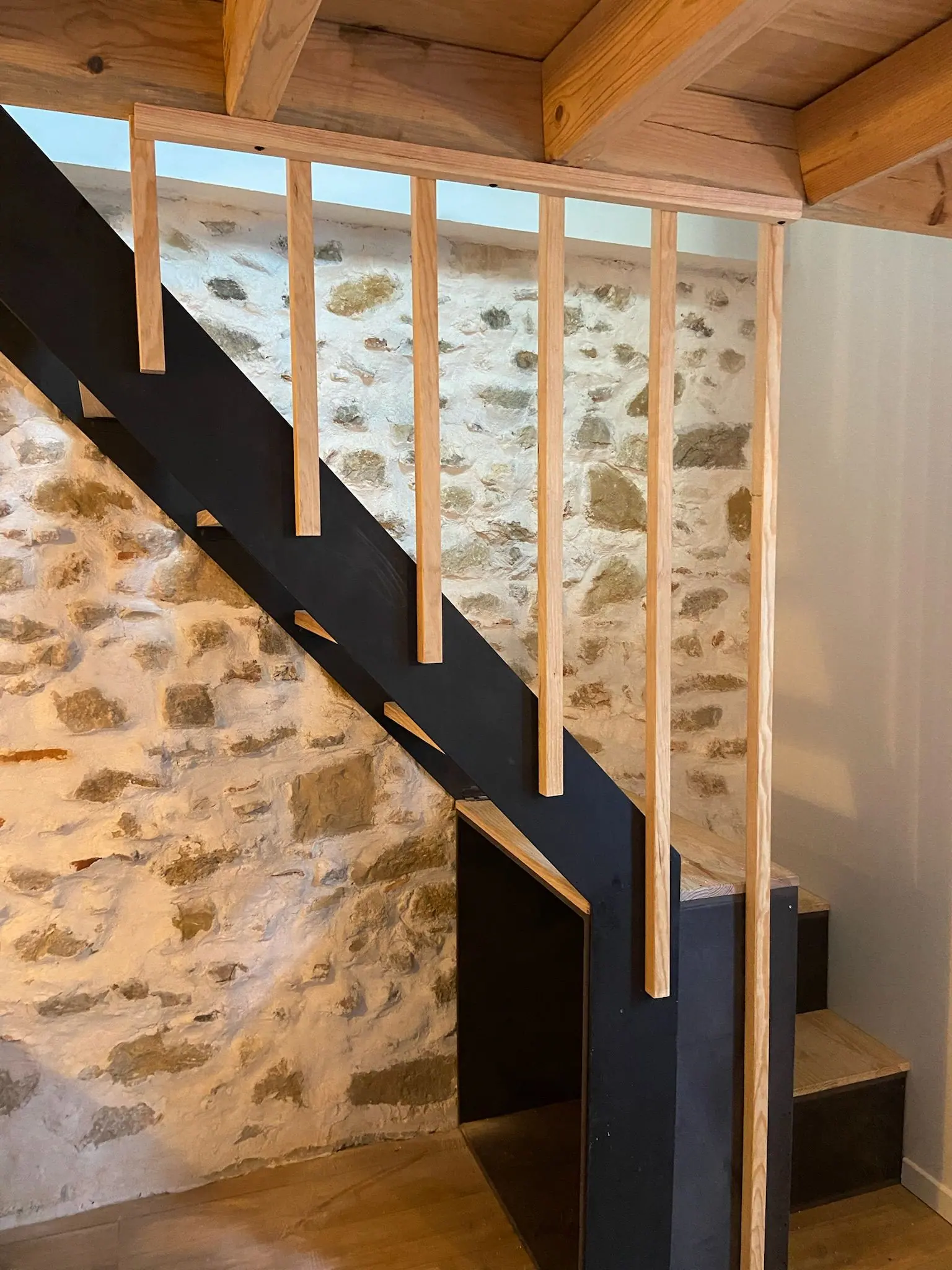 Escalier à limon en acier noir, barreaux en bois et mur de pierre apparente